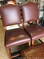 Two brown leather Fremarc Designs bar stools with nail head trim, wooden legs, and padded backrests and seats shown side by side.