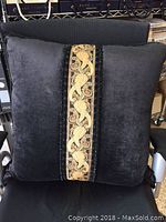 Black velvet pillow with central gold and beige beaded embroidery panel and cording borders, placed on a chair.