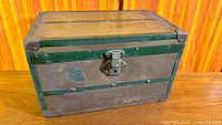 Front view of small antique trunk showing latch, green metal bands, and surface wear