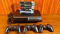 PS3 console with power cord, three controllers and stack of game cases