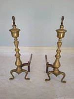 Front view of both brass andirons showing turned columns, finials, cabriole legs and attached iron log rests
