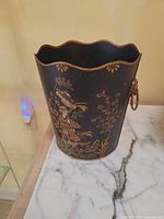 Full view of black painted tin wastebasket showing gold chinoiserie design, scalloped rim, side ring handle, placed on marble surface