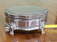 Three-quarter view of sterling silver trinket box showing glass lid, decorative borders, lion feet and measuring tape for scale