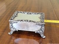 Oblique view of sterling silver footed trinket box showing ornate scrollwork and measuring tape for scale