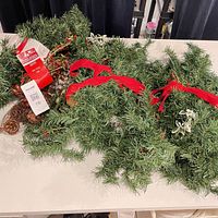 Three green artificial pine garlands on table, two with pinecones/berries, red bow visible