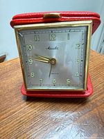 Front view of Mauthe travel clock open in red case showing dial and hands