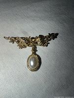 Front view showing ornate gold-tone scroll bar and dangling oval faux pearl