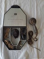 Open Acousticon case showing hearing aid body, cords, charger, earbuds, WWII microphone