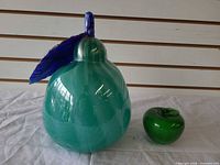 Large green glass pear with blue leaf and stem beside small green apple