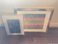 Both framed artworks leaning against wall on hardwood floor, showing relative sizes and frames