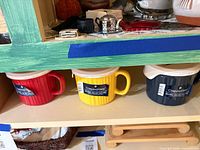 Three CorningWare meal mugs on shelf: red, yellow, navy blue, each with lid
