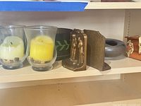 Shelf showing glass hurricane candle holders, brass bookends, black votive, grey tealight holder, wooden box