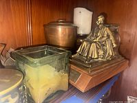 Shelf showing bronze figurine, brass pot, square green glass candle holder, partial view of metal candle holder