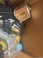 Box of mixed items showing phone accessories, cables, card game box