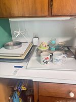 Stack of baking sheets, cake pan, serving tray with mugs, additional small items in background