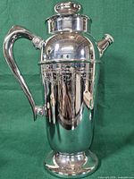 Full view of silver-plated cocktail shaker showing form, handle, spout, pedestal base, engraved grapevine band
