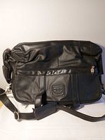 Front view of black leather laptop bag showing handles, shoulder strap, front pocket, logo patch