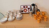 All items on table: mugs, saucers, boxed demitasse set, two moka pots, Gevalia carafe, orange mugs
