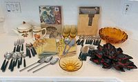 Lot overview displaying all items: tablecloth packages, linens, flatware, amber glass pieces, cookie jar, mug, tumblers