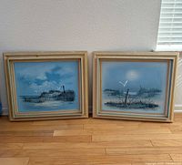 Both framed coastal paintings side by side on floor