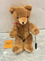 Front view of brown plush teddy bear with red ribbon and size reference pen