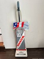Full view gray and white Oreck XL upright vacuum standing against wall with lot tag