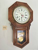 Front view of wooden wall clock showing dial, case shape and lower glass door with pendulum
