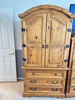 Front view of tall armoire showing doors, drawers, arched top, black metal hardware and honey wood finish