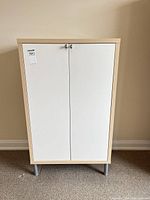 Front view of cabinet showing white doors, light wood frame, metal legs