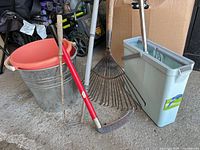 Group shot of all items: metal bucket with orange pail, rake, red-handled hoe, sickle, mop bucket with mop