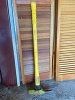 Full view of yellow fiberglass-handle pick mattock leaning against wood paneling