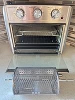 Front view of stainless toaster oven air fryer with door open, wire rack and mesh tray inside, rotisserie drum basket in foreground