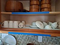 Cabinet shelf with teak bowls on top shelf, ceramic mugs, plates and cups on lower shelf