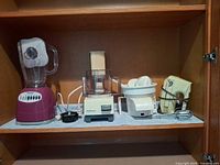 Four small kitchen appliances lined on shelf: blender, food processor, juicer, hand mixer