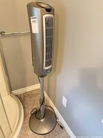 Front view of silver Lasko tower fan on pedestal stand between shower and wall outlet