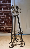 Full view of black metal easel standing, showing scrollwork and leaf details