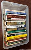 Top view of open clear tote showing approximately 16 paperback books stacked, spines legible