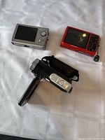 Rear view of silver Olympus camera, red Olympus camera, and black Vivitar camcorder on white surface