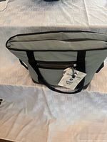 Full view of cooler tote standing upright on table, showing light grey exterior, black straps, front zip pocket, hang tag attached