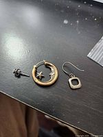 Three single earrings laid out: stud with blue stone, hoop with crescent moon and star, fishhook dangle with square drop