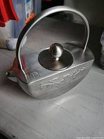Side view of brushed stainless teapot showing embossed motif, handle, lid and spout