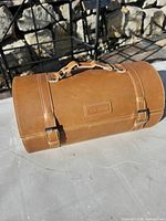 Exterior of brown leather cylindrical case showing handle, buckles, embossed logo