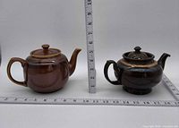 Both teapots side by side on rulers for scale