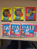 Front view of six sealed ALF trading card/sticker packs: three Topps ALF packs and three Zoot U.S. of ALF 1st Series packs