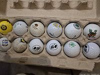 Open egg-carton tray holding 12 logo golf balls, 11 white, 1 yellow, showing various printed course logos