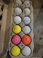 open egg carton showing 12 golf balls, mix of white and colored Pinnacle, Nitro etc.