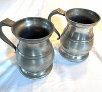 two matching antique pewter pot-bellied pitchers with handles and footed bases on white cloth