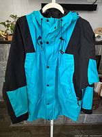 Front view of teal/black hooded rain shell jacket on hanger