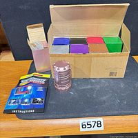 Open shipping box holding eight assorted-color compact lanterns, one bronze unit displayed with handles extended, instruction sheet visible