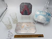Group view of three foil trivets and two etched eagle glasses with ruler for scale